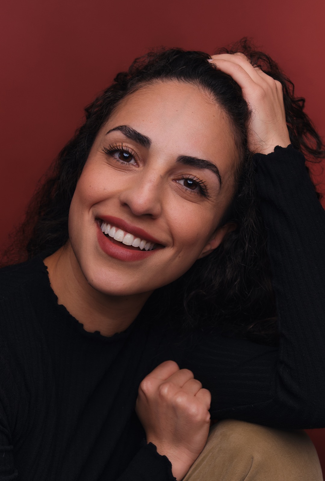 Parmiss Sehat — actor headshot. Warm smile, curly dark hair, against a rich terracotta background.